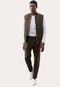 Leichte, gepolsterte olivgrüne Weste über einem weißen Langarmshirt, kombiniert mit dunkelbraunen Hosen und weißen Sneakers. Einfaches Design, keine Muster.