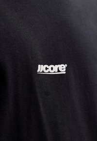 Zwart T-shirt met een geribbelde textuur en een wit geborduurd logo "jjcore" op het linkerborstgedeelte. Simpel en minimalistisch ontwerp.