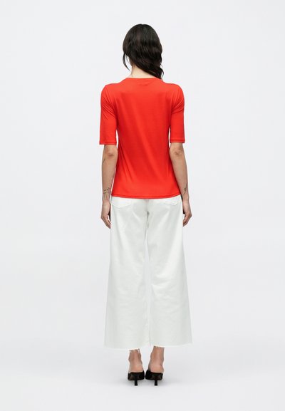 Piros testhez álló rövid ujjú póló fehér, bőszárú nadrággal párosítva. A szövet simának tűnik, enyhe fényességgel és minimális részletekkel.