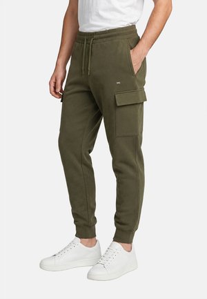 Uomo che indossa pantaloni cargo jogger verde oliva con vita a coulisse e sneakers bianche, in piedi con una mano in tasca.