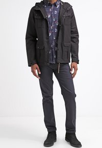 Giacca nera con cappuccio e numerose tasche frontali sopra una camicia a fantasia blu. Pantaloni grigi e stivali neri completano l'outfit.