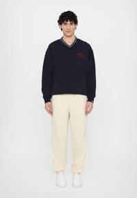 Sweatshirt azul marinho com texto vermelho "GYMNASIUM", decote em V e punhos canelados, combinado com calças de moletom creme e ténis brancos.