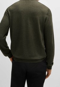 Mężczyzna w oliwkowej zielonej bluzie z długim rękawem i czarnych spodniach, widziany z tyłu na jednolitym tle.