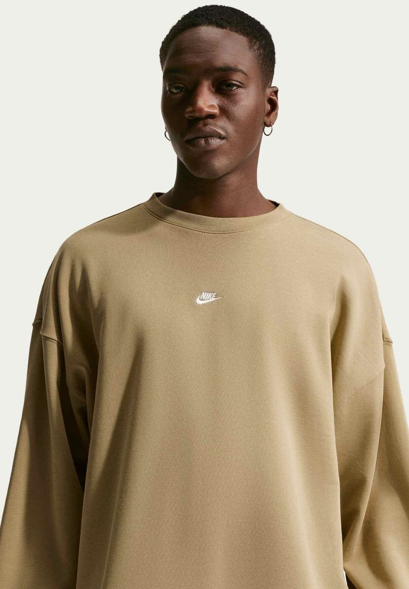 Giovane uomo con capelli corti e orecchini a cerchio che indossa una felpa Nike beige larga su uno sfondo chiaro.
