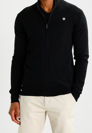 Mann, der einen schwarzen Reißverschluss-Pullover mit kleinem TS-Logo auf der Brust und beige Hose trägt, steht mit teilweise in die Taschen gesteckten Händen.