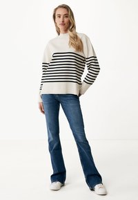 Pull en maille blanc à rayures horizontales noires, associé à un jean bleu évasé et des baskets blanches. Le pull présente un col montant et une coupe décontractée.