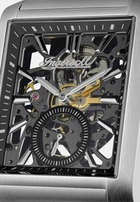 Srebrny zegarek ze stali nierdzewnej z czaszą typu skeleton, ozdobioną złotymi i czarnymi akcentami, wyraźnymi znacznikami godzin oraz dekoracyjnym subtarczą.