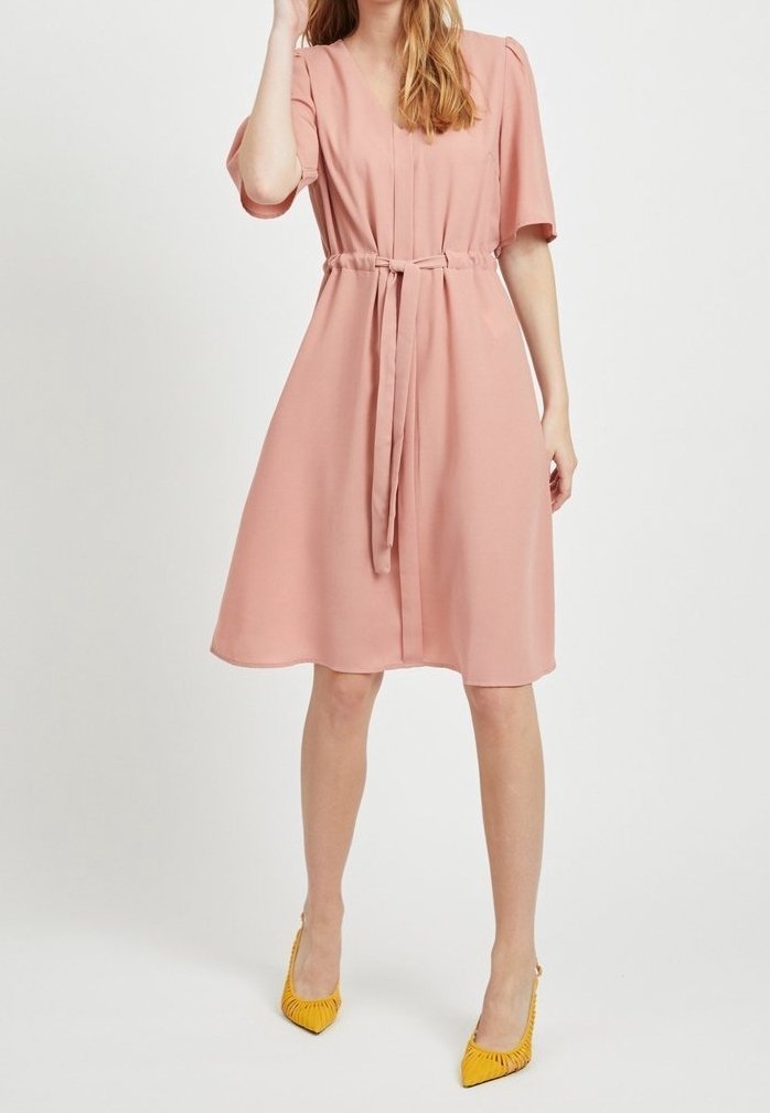 Femme portant une robe rose clair au genou avec des manches courtes et une ceinture à la taille, associée à des talons hauts jaunes à bout pointu.