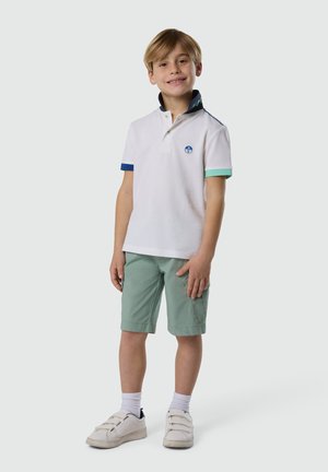 Niño sonriente que lleva un polo blanco con mangas azules y verdes, pantalones cortos verdes, calcetines blancos y zapatillas blancas, posando frente a un fondo liso.