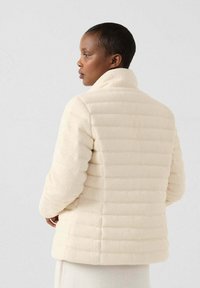 Chaqueta acolchada color crema que presenta una textura suave, cuello alto y un diseño estructurado con paneles horizontales. La vista trasera muestra una silueta elegante.