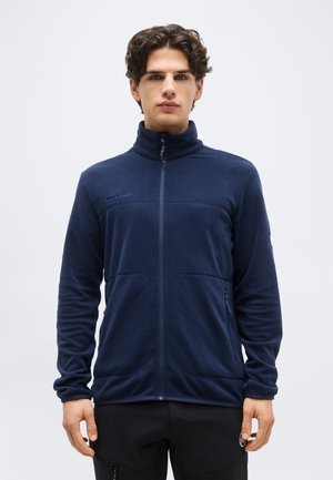 Mladić s kovrčavom kosom odjeven u tamnoplavu flis jaknu na patentni zatvarač i crne hlače, stoji na jednobojnoj svijetloj pozadini.