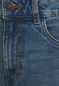 Denimstoff in Mittelblau mit subtiler Ausbleichung. Mit orangen Nähten, einem oberen Knopf und einer Seitentasche. Nahaufnahme der Stoffstruktur.