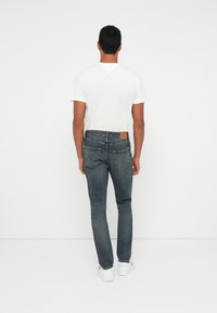 Mees, kelles on valge T-särk, sinised teksad ja valged tossud, seisab valge seina poole pöördunult, käed lõdvestunult külgedel.