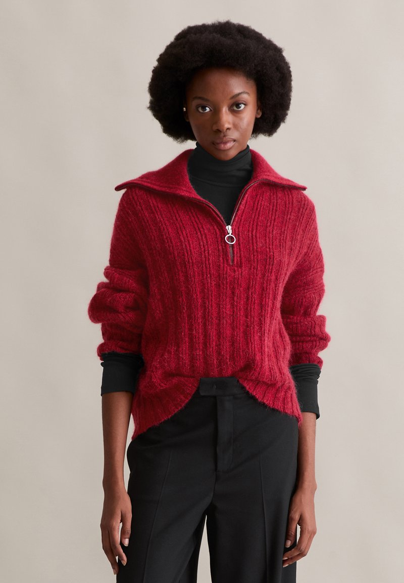 Roter gestrickter Pullover mit einem Reißverschluss und hohem Kragen, gerippter Textur und weiten Ärmeln. Über einem schwarzen Rollkragenpullover getragen und mit schwarzen Hosen kombiniert.