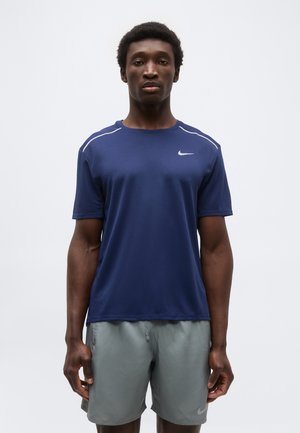 Άνδρας στέκεται με πρόσωπο προς τα εμπρός, φορώντας μπλε σκούρο αθλητικό μπλουζάκι με κοντά μανίκια και ανοιχτό γκρι αθλητικό σορτς, με φόντο ένα απλό ανοιχτόχρωμο υπόβαθρο.