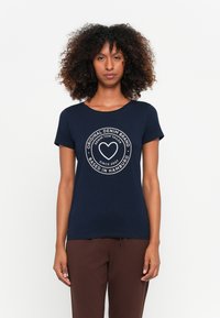 Mulher com cabelo encaracolado vestindo uma t-shirt azul marinho com um logótipo circular branco e texto, combinada com calças castanhas, de pé contra uma parede branca.