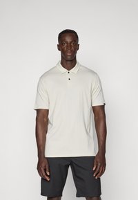 Polo-shirt beige clair avec col, deux boutons noirs, manches courtes et une étiquette sur la manche gauche ; associé à un short noir.