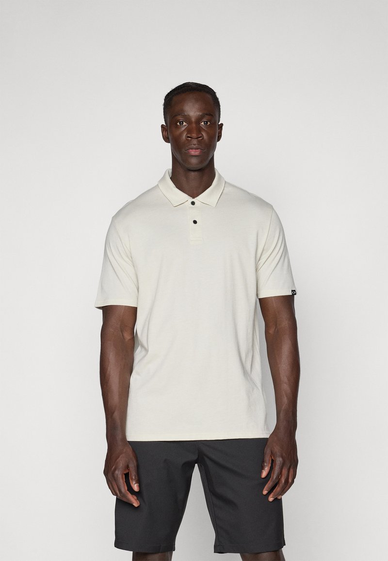 Polo-shirt beige clair avec col, deux boutons noirs, manches courtes et une étiquette sur la manche gauche ; associé à un short noir.