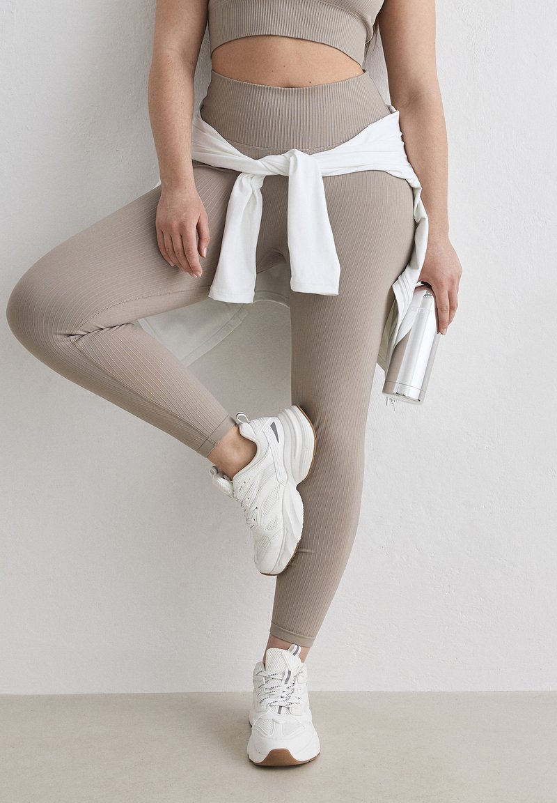 Persona con leggings beige acanalados y top corto, zapatillas blancas y una sudadera blanca atada a la cintura, sosteniendo una botella de agua plateada.