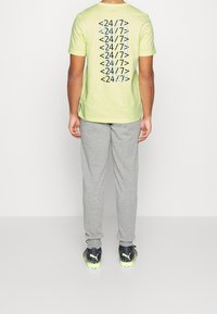 Camiseta verde claro con patrón de texto negro "<24/7>" en la parte posterior, combinada con pantalones de chándal grises y zapatos negros con acentos verdes.