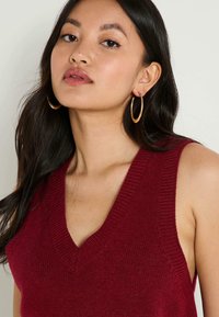 Jeune femme aux cheveux bruns portant des boucles d'oreilles en or et un haut sans manches en tricot bordeaux à décolleté en V.