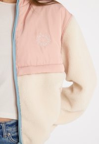 Polares és nylon dzseki, pink felső résszel és krémszínű alsó résszel. Hímzett napmotívummal és világoskék cipzárral rendelkezik.