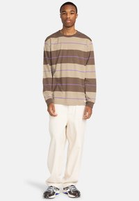 Langeærmet stribet skjorte i brun og beige med lilla detaljer, udstyret med en brystlomme; båret med cremede bukser og sneakers.