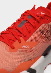 Κοντινό πλάνο φωτεινού κόκκινου και πορτοκαλί αθλητικού παπουτσιού με χοντρές κορδόνια, πλέγμα στο πάνω μέρος, ετικέτα "PRO 3" και σήμανση "The North Face" στο πλάι.