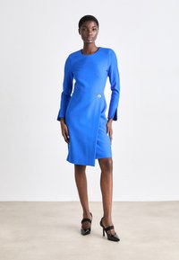 Μπλε, μακρυμάνικο φόρεμα με στρογγυλή λαιμόκοψη, πλάγιο σκίσιμο και λεπτομέρεια με ασημένιο κουμπί. Απαλή ύφανση; το μοντέλο το έχει συνδυάσει με μαύρες μυτερές γόβες.