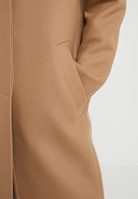 Personne portant un manteau couleur camel avec la main droite dans la poche, vue de l'épaule à la cuisse sur un fond clair et uni.