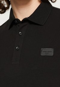 Černé polo tričko s klasickým límečkem, dvěma knoflíky, texturovanou tkaninou a pravoúhlou nášivkou na prsou.