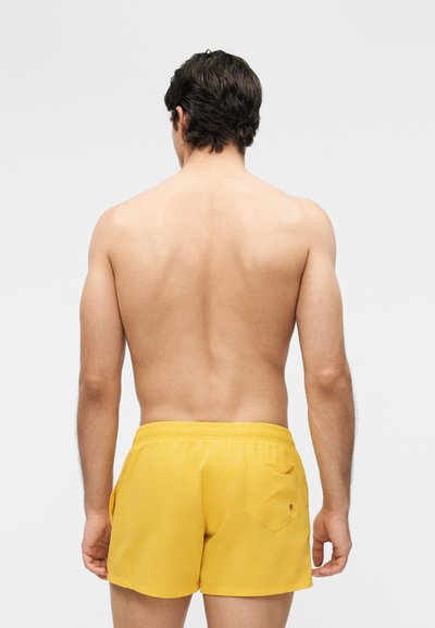 Άνδρας με σκούρα μαλλιά στέκεται με την πλάτη προς τα εμπρός, φοράει κίτρινα μαγιό με τσέπη, σε μια απλή λευκή φόντο.