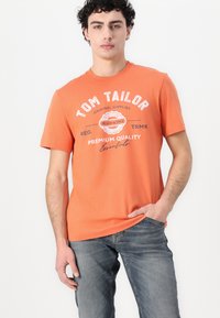 Oranžs kokvilnas T-krekls ar apaļu kaklu, īsām piedurknēm un baltu drukātu tekstu ar apļa logotipu. Valkāts kopā ar ziliem džinsu biksēm.