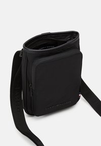 Sort nylon crossover taske med lynlåse top, frontlomme og justerbar rem. Har prægede logo og kontrastfarvet detalje nær remmen.