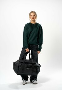Borsa a sacca nera in materiale resistente, con più manici e un compartimento con zip. Modello che indossa una felpa verde e jeans scuri.