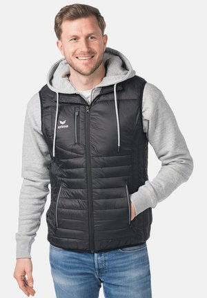 Smiling man wearing a black quilted vest over a grey hoodie with hands in pockets, standing against a plain light background.