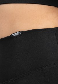 Leggings neri con elastico in vita, dalla texture liscia, con un piccolo etichetta del marchio "SMILOOX" in vita e cuciture piatte.