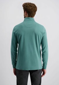 Mintgroene pullover met lange mouwen en hoge kraag, gladde textuur en een subtiele naaddetail op de rug. Gecombineerd met donkere broeken.