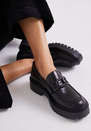 Sort chunky loafer med tyk sål og kædedetalje båret med sorte cropped bukser på krydsede ben mod hvid baggrund.