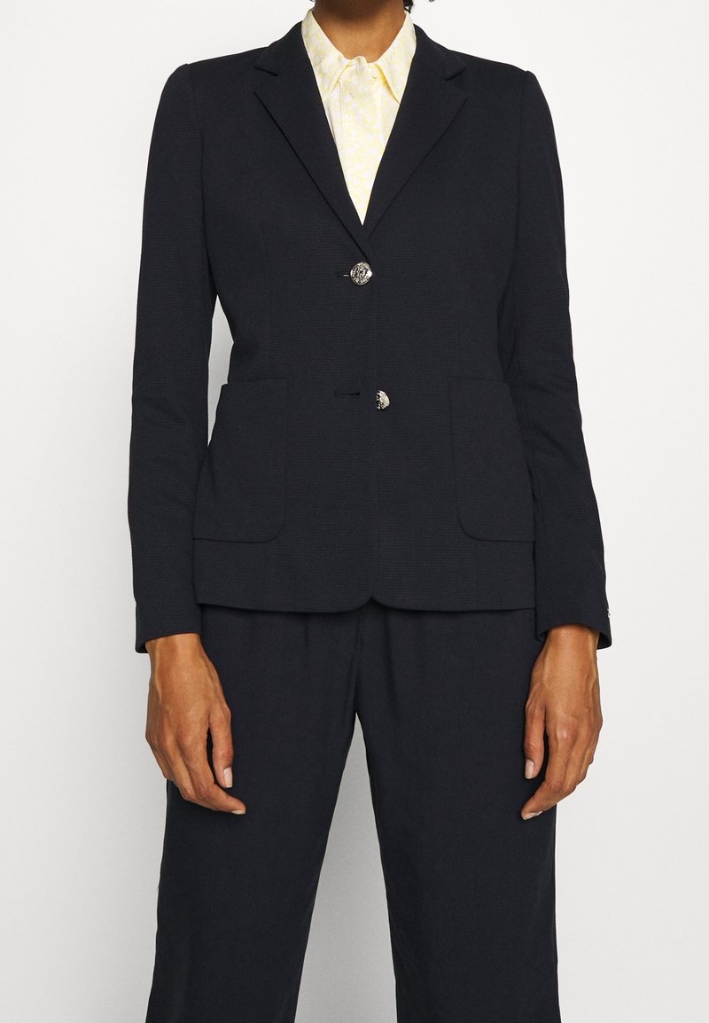 Femme portant un blazer noir sur mesure avec des boutons argentés sur une chemise jaune clair à motifs, assorti d’un pantalon noir à jambes droites.