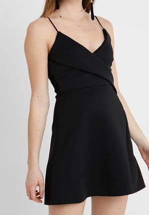 Kobieta ubrana w czarną sukienkę koktajlową bez rękawów z cienkimi ramiączkami, dekoltem w serek i kopertowym stylem gorsetu, na jednolitym tle.