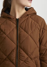 Veste matelassée brune avec capuche, présentant une fermeture éclair noire, un motif en losanges et des détails de couture visibles. Tissu doux et léger.