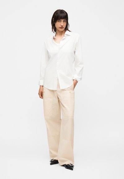 Γυναίκα με σκουρόχρωμα μαλλιά που φοράει λευκό πουκάμισο με κουμπιά, μπεζ φαρδιά παντελόνια και μαύρα μυτερά παπούτσια, στέκεται μπροστά σε ένα απλό λευκό φόντο.