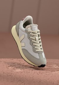 Sneaker con tomaia in rete bianca con accenti in suede grigio, lacci crema e suola gialla testurizzata. Include il logo "VEJA" sul tallone.