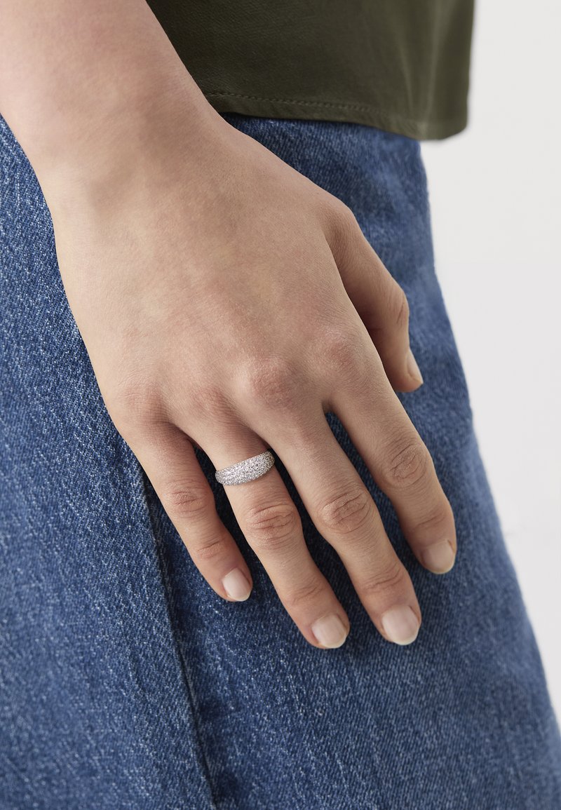 Main reposant sur un jean en denim bleu, portant une bague en argent incrustée de petites pierres brillantes à l'annulaire.