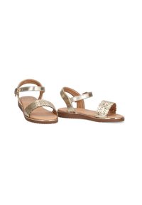 Sandalias metálicas doradas con una amplia tira que presenta un patrón floral cortado a láser, tira adjustable en el tobillo y suela plana marrón.