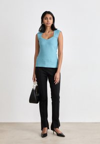 Top in cotone turchese a coste con scollatura ampia e forma aderente, abbinato a pantaloni neri su misura e décolleté a punta; tiene in mano una borsa nera.