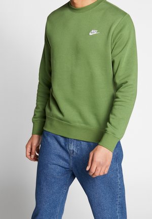 Mężczyzna ubrany w oliwkową bluzę Nike z logo na piersi i niebieskie dżinsy, stojący na białym tle.