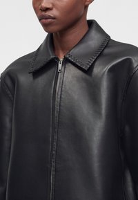 Gros plan d'une personne portant une veste en cuir noire avec une fermeture éclair à l'avant et des coutures détaillées au niveau du col.