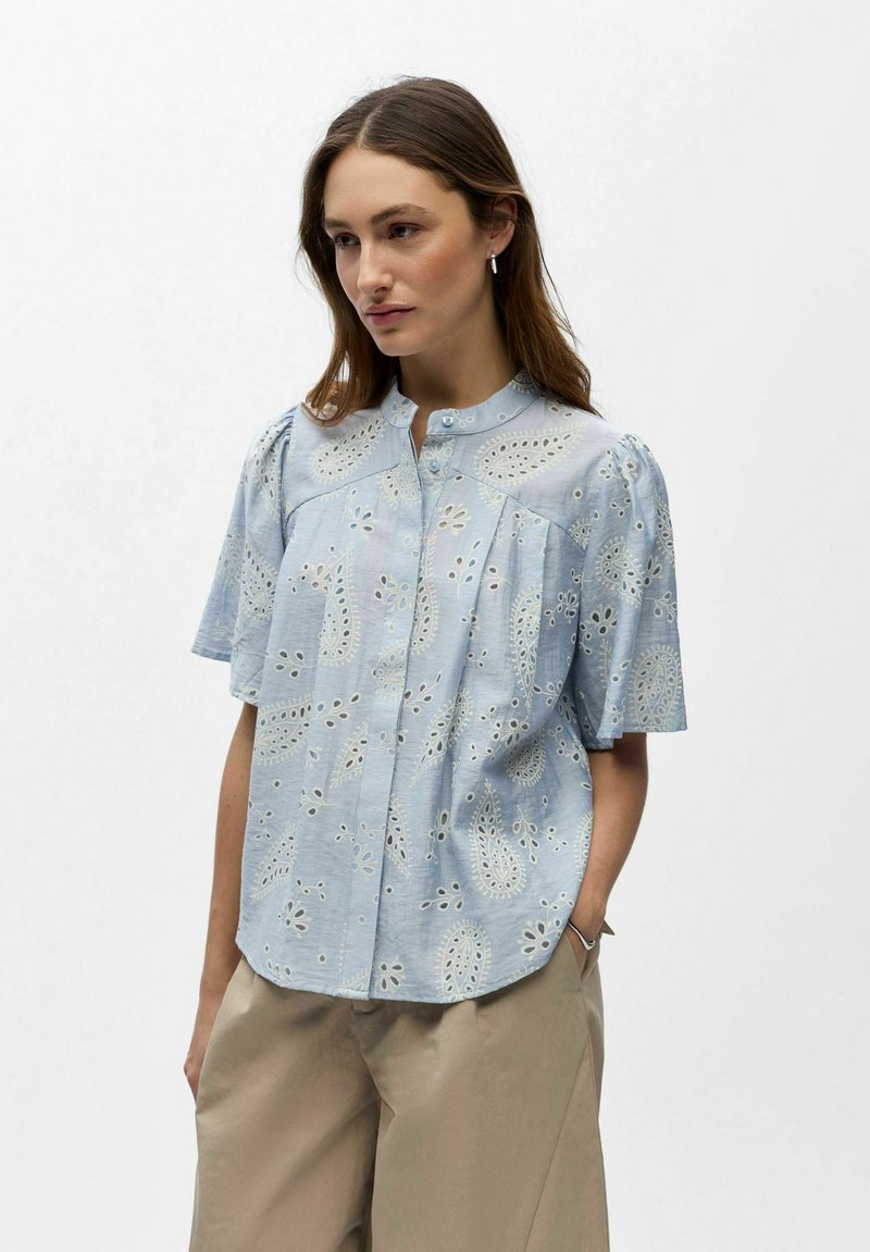 Femme portant un chemisier à manches courtes bleu clair avec des motifs paisley en broderie anglaise blanche et un pantalon beige, debout avec une main dans la poche.